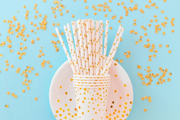 Birthday or party paper glasses with straws, dish and golden sequins on blue background. Festive concept. Flat lay style. Top view.