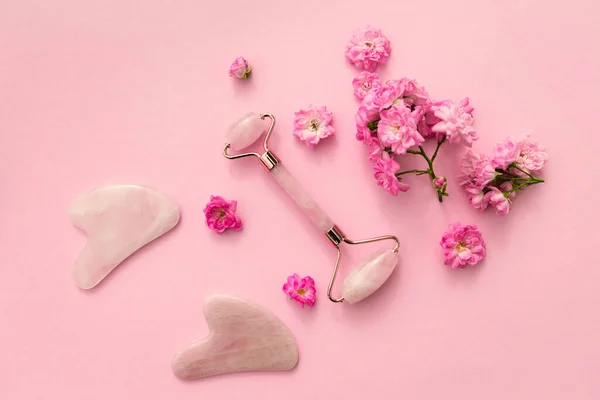 Ev spası için yüz masajı seti. Gül tomurcuklarıyla pembe pastel arka planda gül kuvarsından yapılmış yüz silindiri ve gua sha masaj aleti. Doğal tedavi kavramı.
