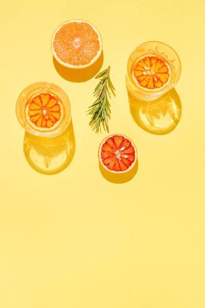 Yarım greyfurt ve kanlı portakal, biberiye ve parlak sarı zemin üzerinde su olan iki bardak. Yaz tatili konsepti. Düz güneş ışığı yatıyordu. Asgari düzeyde. Üst Manzara. Boşluğu kopyala