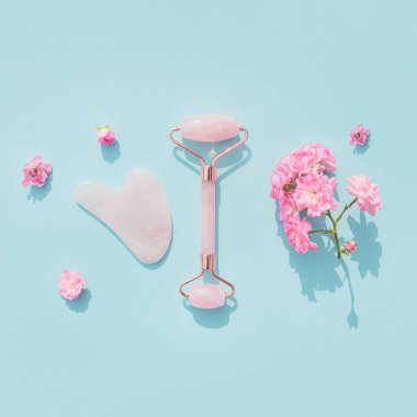 Ev spası için yüz masajı seti. Gül tomurcuklarıyla mavi pastel arka planda gül kuvarsından yapılmış yüz silindiri ve gua sha masaj aleti. Doğal tedavi konsepti. Düz yatıyordu. Üst görünüm