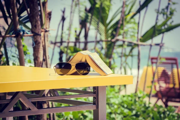 Libro y gafas de sol tumbados sobre una mesa amarilla en una playa tropical —  Fotos de Stock