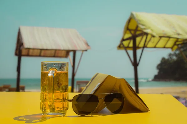 Copo de cerveja, óculos de sol e um livro em uma mesa amarela em uma bea — Fotografia de Stock