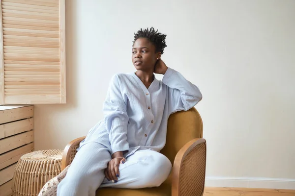 stock image Young girl in the morning in a cozy blue pajamas sits in a brown armchair and looks out the window.