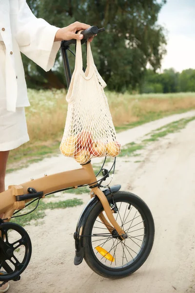 String bag met appels weegt op het stuur van een vouwfiets. Vervolgens een meisje in wit linnen pak. Het concept van een gezonde levensstijl en het gebruik van herbruikbare milieuvriendelijke dingen. — Stockfoto