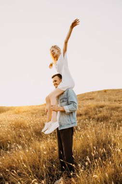 Güzel genç bir kadın gün batımında erkek arkadaşının omuzlarında çayırda