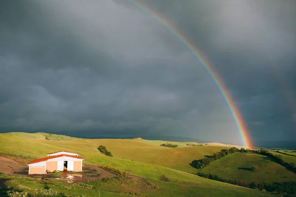scenic shot of stormy clouds and rainbow over green meadow and house