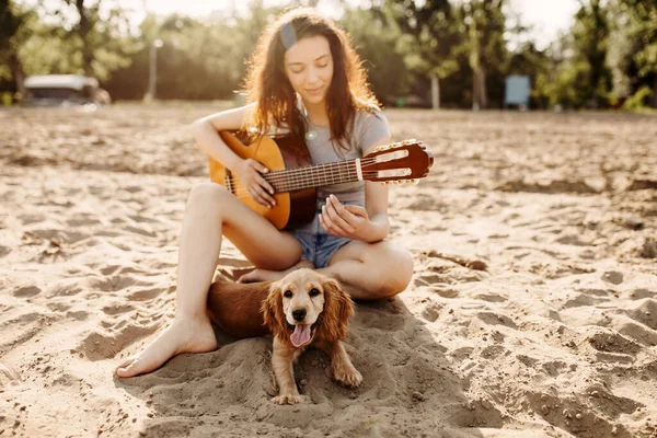 一位年轻貌美的女士一边弹吉他 一边和她的小猎犬在沙滩上玩耍 — 图库照片