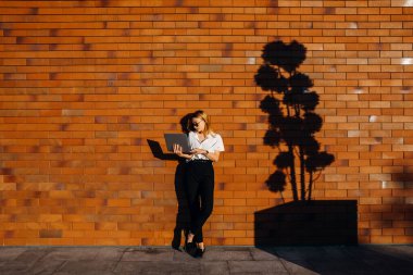 Kırmızı tuğla duvarın önünde dizüstü bilgisayarla çalışan güzel genç bir kadın.