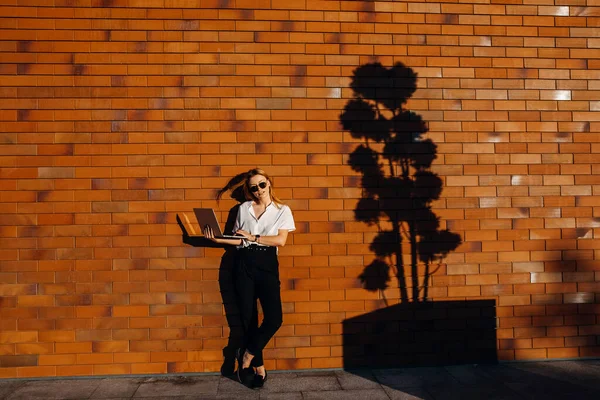 Kırmızı tuğla duvarın önünde dizüstü bilgisayarla çalışan güzel genç bir kadın.