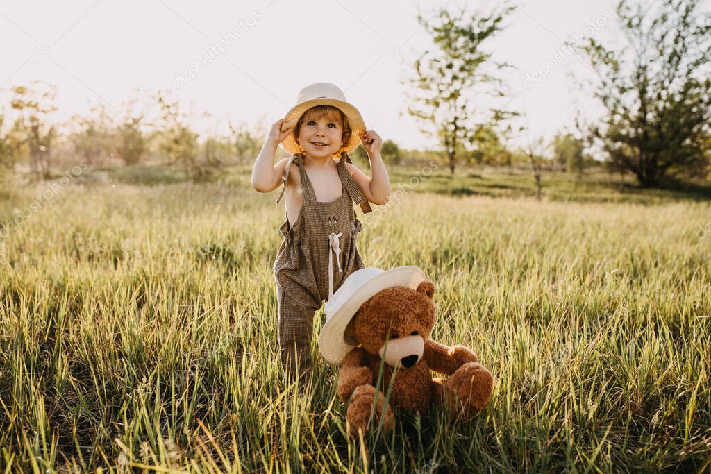 adorable little boy in stylish clothes and straw hat on nature with his teddy bear