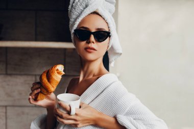 Ayçöreği ve bir fincan kahveyle havluyla ve bornozuyla otelde dinlenen güzel genç bir kadın.