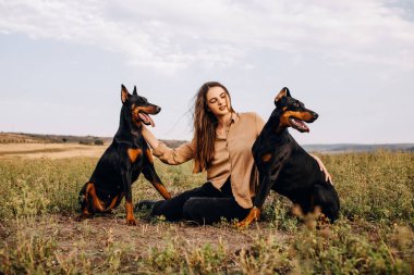 Doğada oturan iki Doberman köpeğiyle güzel bir kadının portresi.