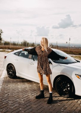 Güzel genç bir kadın arabasının önünde duruyor.