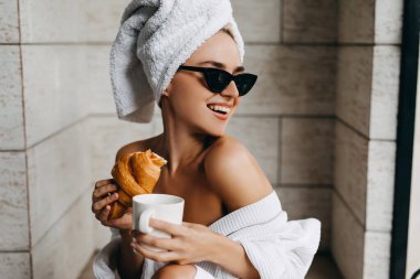 Ayçöreği ve bir fincan kahveyle havluyla ve bornozuyla otelde dinlenen güzel genç bir kadın.