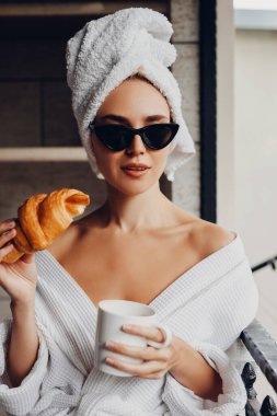 Ayçöreği ve bir fincan kahveyle havluyla ve bornozuyla otelde dinlenen güzel genç bir kadın.