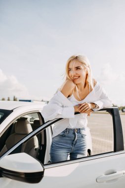 Güzel genç bir kadın arabasının önünde duruyor.