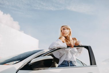 Güzel genç bir kadın arabasının önünde duruyor.