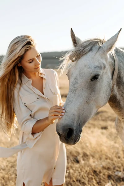 portrait of beautiful blonde woman with white horse on nature during sunset