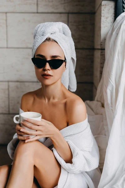 白いバスローブにタオルを頭にサングラスをかけコーヒーを飲みながら家でリラックスした美しい若い女性 — ストック写真