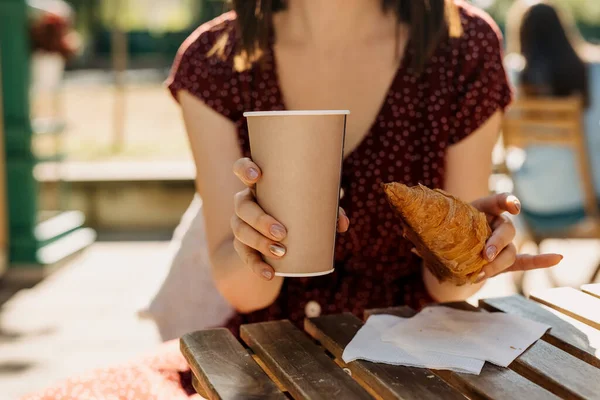 ホットドリンクとクロワッサンの紙コップと一緒にストリートカフェに座っている女性のクロップドショット — ストック写真