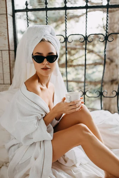 白いバスローブにタオルを頭にサングラスをかけコーヒーを飲みながら家でリラックスした美しい若い女性 — ストック写真