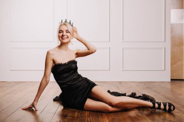 Taçlı, siyah elbiseli, gülümseyen, yerde oturan mutlu, genç sarışın kadın..