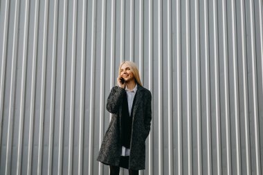 Metalik gri bir duvarın karşısında duran şık, genç bir kadın akıllı telefonda konuşuyor..