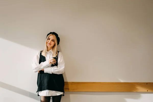 年轻时髦的金发女人靠着白墙站着 手里拿着一杯咖啡 面带微笑 — 图库照片