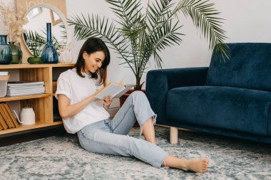 Evde kitap okuyan, oturma odasında yerde oturan mutlu genç bir kadın..