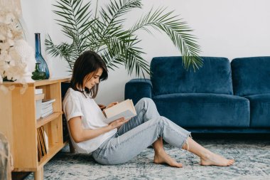 Genç esmer kadın evde kitap okuyor, oturma odasında yerde oturuyor..