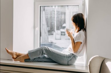 Pencerenin kenarında oturan genç bir kadın, elinde beyaz bir fincan kahve tutuyor..