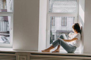 Güneş ışığında pencere kenarında oturan genç bir kadın elinde bir fincan kahve tutuyor..