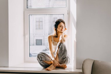 Güneş ışığında pencere kenarında oturan genç bir kadın, kablosuz kulaklıkla müzik dinliyor..