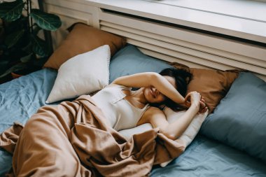 Genç bir kadın sabah yatağa uzanmış, gülümsüyor, elleriyle yüzünü örtüyor..