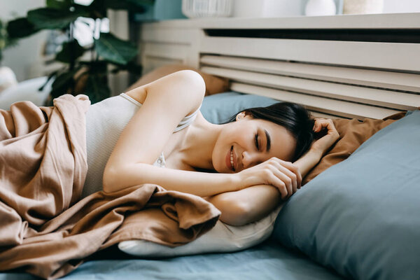 Молодая женщина лежит в постели утром, улыбаясь, с закрытыми глазами.