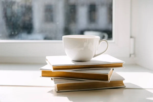 Blank white cup of coffee or tea on books by the window in sunlight.