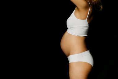 Beyaz spor iç çamaşırı giyen hamile bir kadın. Siyah arka planda doğrudan güneş ışığı altında. Yakın plan..