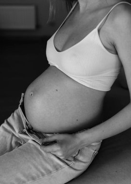 Hamile bir kadın evdeki kanepede oturuyor. Kırpılmış bluz ve kot pantolon giyiyor. Yakın plan..