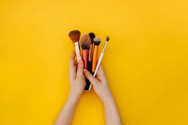 Sarı arka planda çeşitli makyaj fırçaları tutan kadın eller..