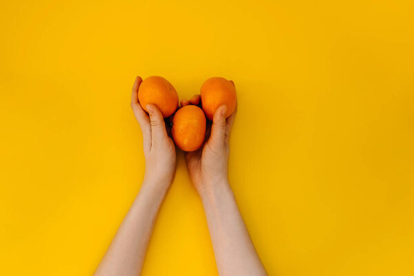 Female hands holding three clementines on yellow background.