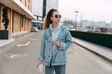 Yeniden kullanılabilir bir fincan kahvesi olan bir kız, kot ceket giyiyor, açık havada, şehir sokaklarında.