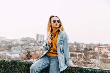 Kot pantolon giyen genç bir kadın, şehrin arka planında yüksek bir yerde çitin üzerinde oturuyor..