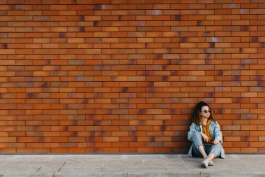 Kot pantolon giyen genç bir kadın, dışarıda tuğla duvar arkasında oturuyor..