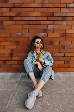 Kot elbiseli ve güneş gözlüklü genç bir kadın, tuğla duvarın yanında oturuyor, açık havada..