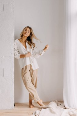 Genç sarışın kadın saçlarıyla oynuyor, beyaz bir gömlek ve pantolon giyiyor, içeride..