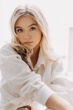 Uzun dalgalı saçlı, beyaz gömlekli, yumuşak pencere ışığında oturan genç sarışın kadın..