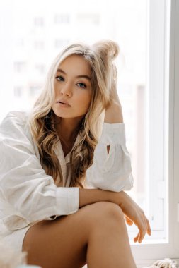 Uzun dalgalı saçlı, beyaz gömlekli, yumuşak pencere ışığında oturan genç sarışın kadın..