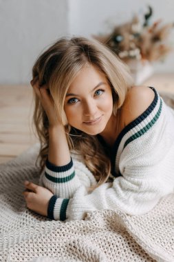 Genç sarışın bir kadının portresi, karnının üstüne yatmış, içerde, kameraya bakıyor, gülümsüyor..