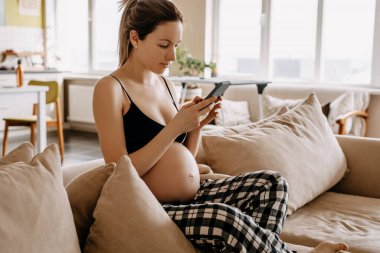 Hamile bir kadın evdeki kanepede oturuyor, mesaj atıyor, akıllı telefon tutuyor..