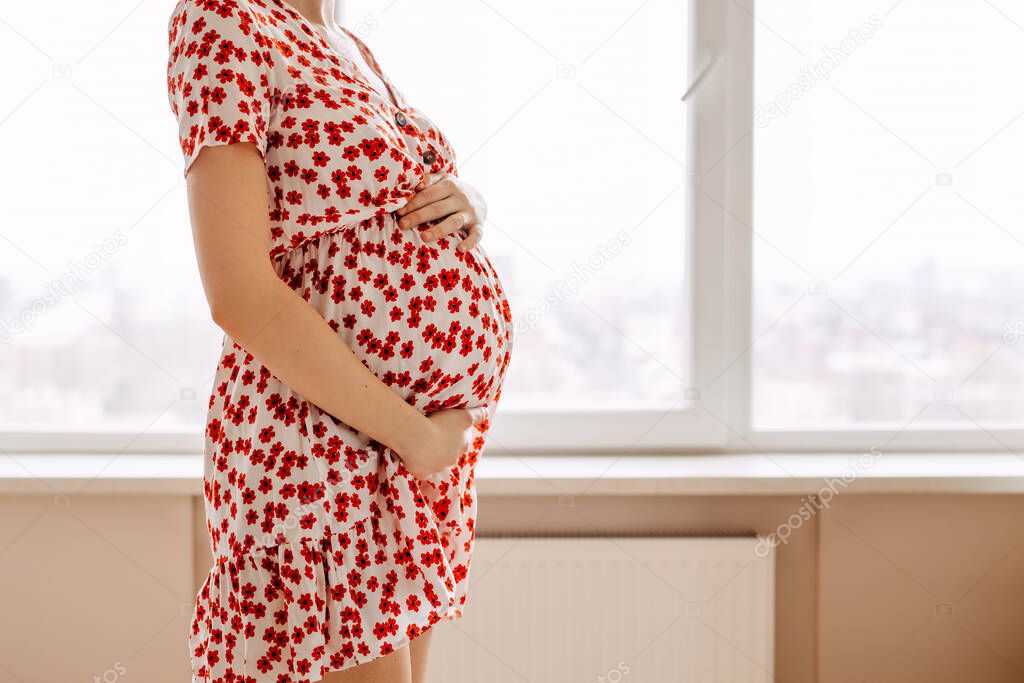 Closeup of a pregnant woman wearing a summer dress, holding hands on belly.